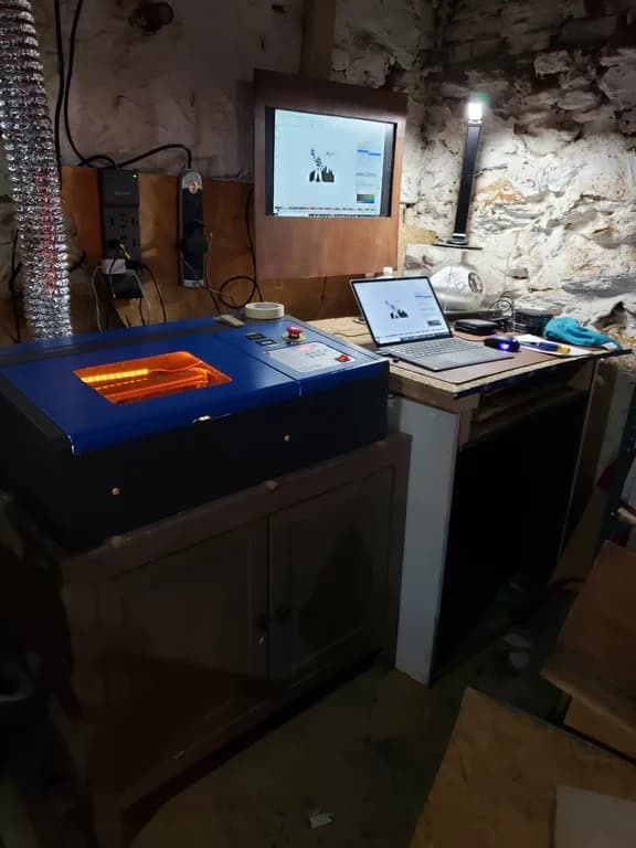 My blue K40 laser in the old stone-walled basement workshop, with its orange-lit lid, wall monitor and laptop.