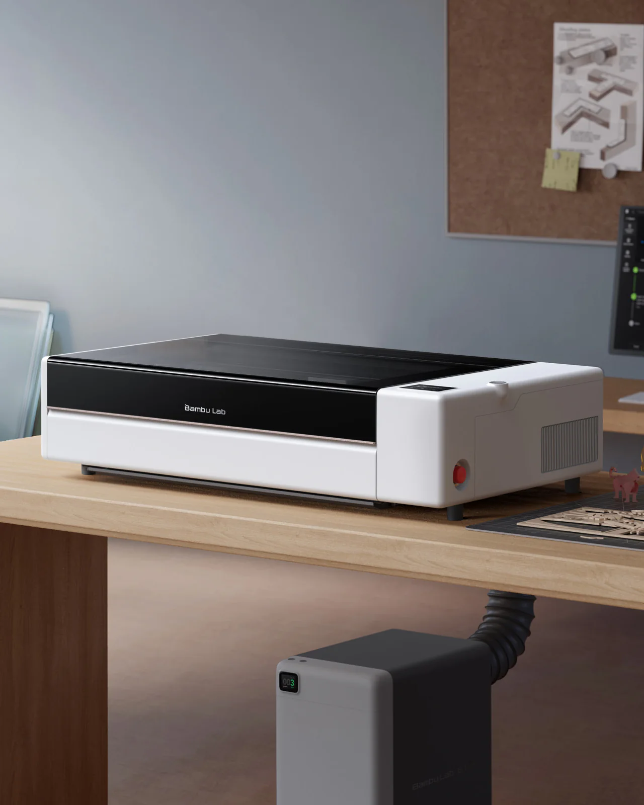 Bambu Lab R1 laser cutter on a workshop desk beside a monitor and 3D-printed parts, with an air purifier connected by a hose on the floor.